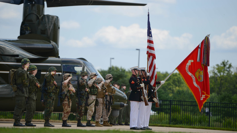 Marines in green and khaki uniforms and helmets salute in front of a helicopter and stand behind a Marine color guard with rifles, an American flag and a Marine Corps flag lowered.