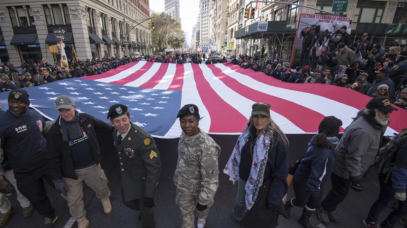 Soldiers in uniform and members of the public carry a huge American flag at shoulder-height in a parade on the streets of New York City. Vietnam veterans are in the background.