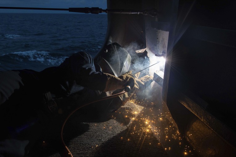 A sailor lies on a ship's deck to weld something through a panel at night.