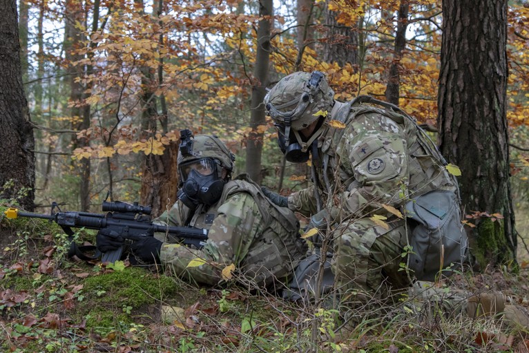 A soldier kneels down next to another soldier on the ground with his weapon pointed.